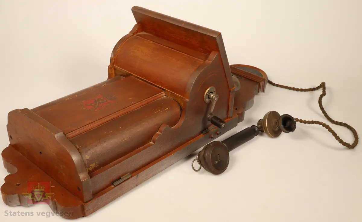Telefonapparat festet til en treplate som er ment til å henges opp på veggen. Treplaten er brunbeist. Telefonapparatet har to bjeller på. Sort og blankt telefonrør. Sveivehåndtak på siden. Den nedre delen av apparatet er preget med våpenskjold og navn på produsent. 