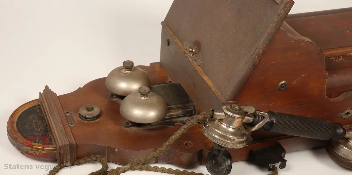 Telefonapparat festet til en treplate som er ment til å henges opp på veggen. Treplaten er brunbeist. Telefonapparatet har to bjeller på. Sort og blankt telefonrør. Sveivehåndtak på siden. Den nedre delen av apparatet er preget med våpenskjold og navn på produsent. 