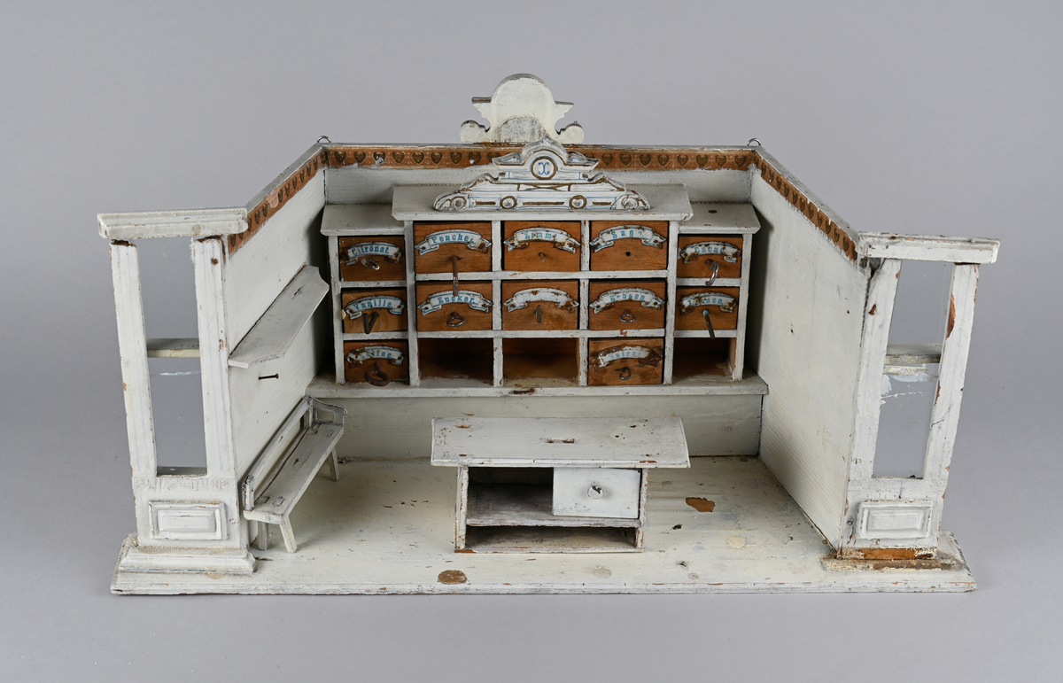 Lekebutikk i tre. Hvitmalt innredning med palmettbord og to utskårne ornamenter øverst. Tre hyllerader med 12 skuffer. Skuffene av ubehandlet tre og navneskilt i metall. To smale glassvegger ytterst. En løs benk og en løs disk med en skuff.