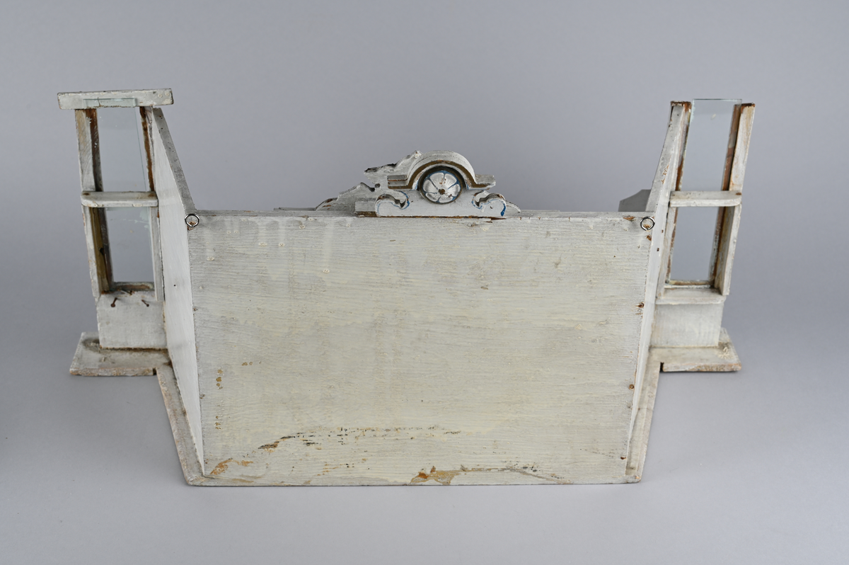 Lekebutikk i tre. Hvitmalt innredning med palmettbord og to utskårne ornamenter øverst. Tre hyllerader med 12 skuffer. Skuffene av ubehandlet tre og navneskilt i metall. To smale glassvegger ytterst. En løs benk og en løs disk med en skuff.