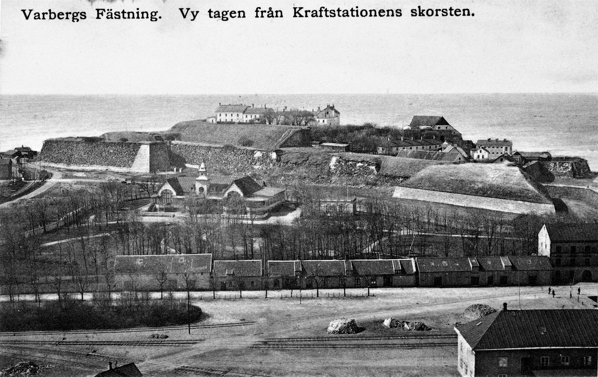Vykort, "Varbergs fästning. Vy tagen från kraftstationens skorsten. I förgrunden ses de låga husen längs Sjöallén framför Societetshuset och parken. Till höger, höljd i träd, står vattenpaviljongen där kurgästerna intog det järnhaltiga brunnsvattnet. Byggnaden revs 1939. Fototid ca 1915.