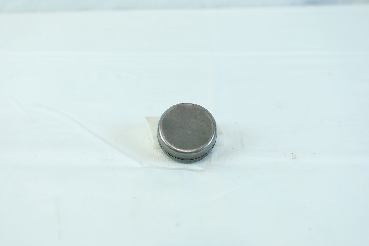 Etui i metall med rund form. I etuiet ligger det noen mindre metallstenger og flere små kors.
