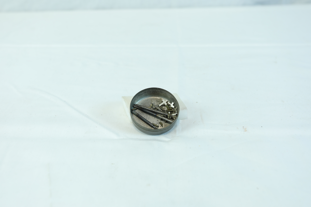 Etui i metall med rund form. I etuiet ligger det noen mindre metallstenger og flere små kors.