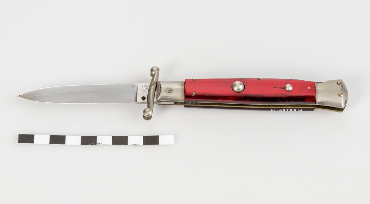 Stilettkniv. Italiensk. Knivblad i bajonettstil, fälls ut åt sidan med tryckknappssystem. I ihopfällt läge finns en säkerhetsspärr. Rött knivskaft i snäckskalsimitation.