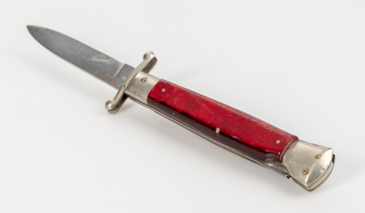 Stilettkniv. Italiensk. Knivblad i bajonettstil, fälls ut åt sidan med tryckknappssystem. I ihopfällt läge finns en säkerhetsspärr. Rött knivskaft i snäckskalsimitation.