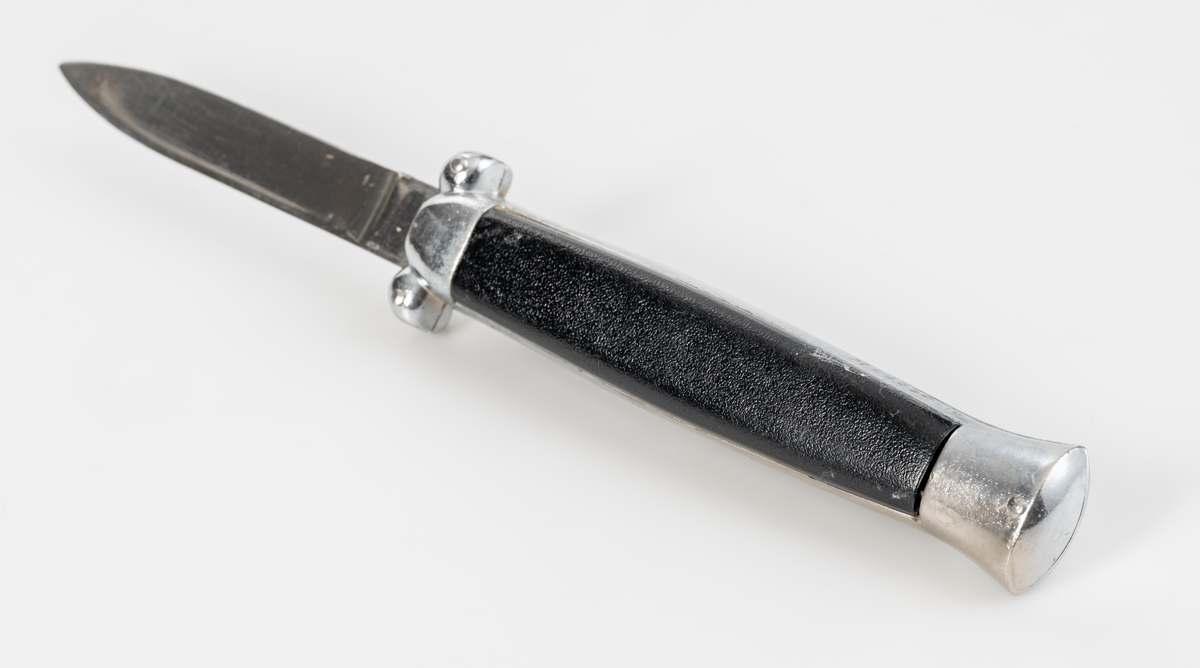 Stilettkniv. Knivblad i bajonettstil som frigörs framåt med skjutknappssystem. Svart knivskaft i plast.