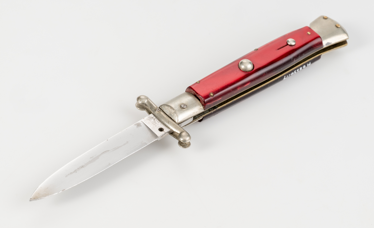 Stilettkniv. Italiensk. Knivblad i bajonettstil, fälls ut åt sidan med tryckknappssystem. I ihopfällt läge finns en säkerhetsspärr. Rött knivskaft i snäckskalsimitation.