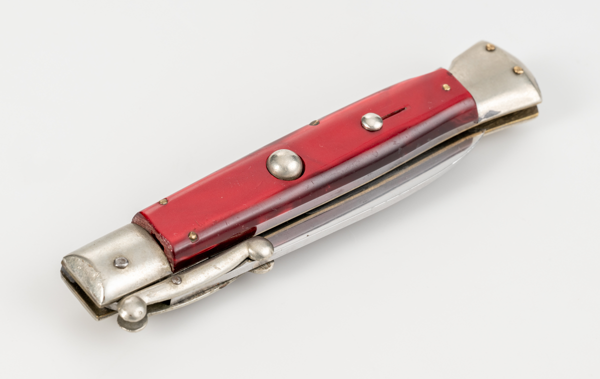 Stilettkniv. Italiensk. Knivblad i bajonettstil, fälls ut åt sidan med tryckknappssystem. I ihopfällt läge finns en säkerhetsspärr. Rött knivskaft i snäckskalsimitation.