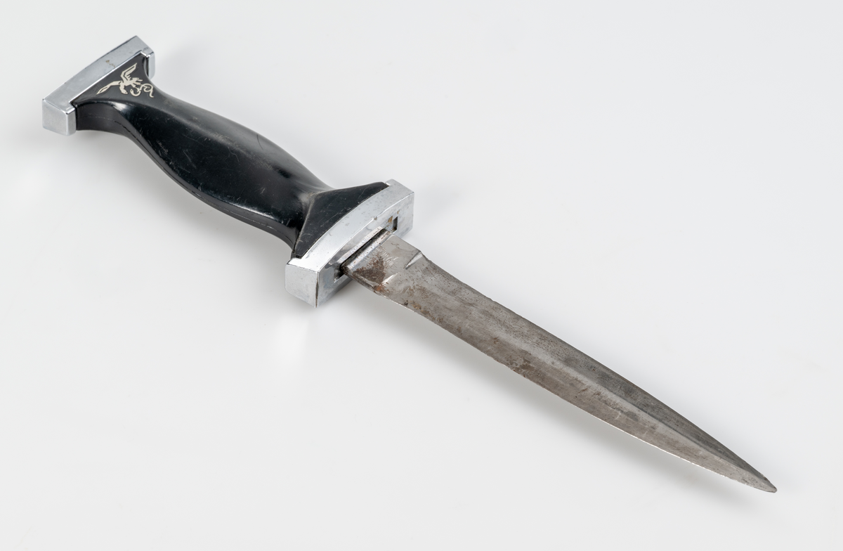 Dolk. Symetrisk med dubbeleggat stålblad. Förnicklad parerstång och profilerat knivskaft i svart plast. Silverfärgad symbol föreställande en örn och en orm på skaftets ena sida.
