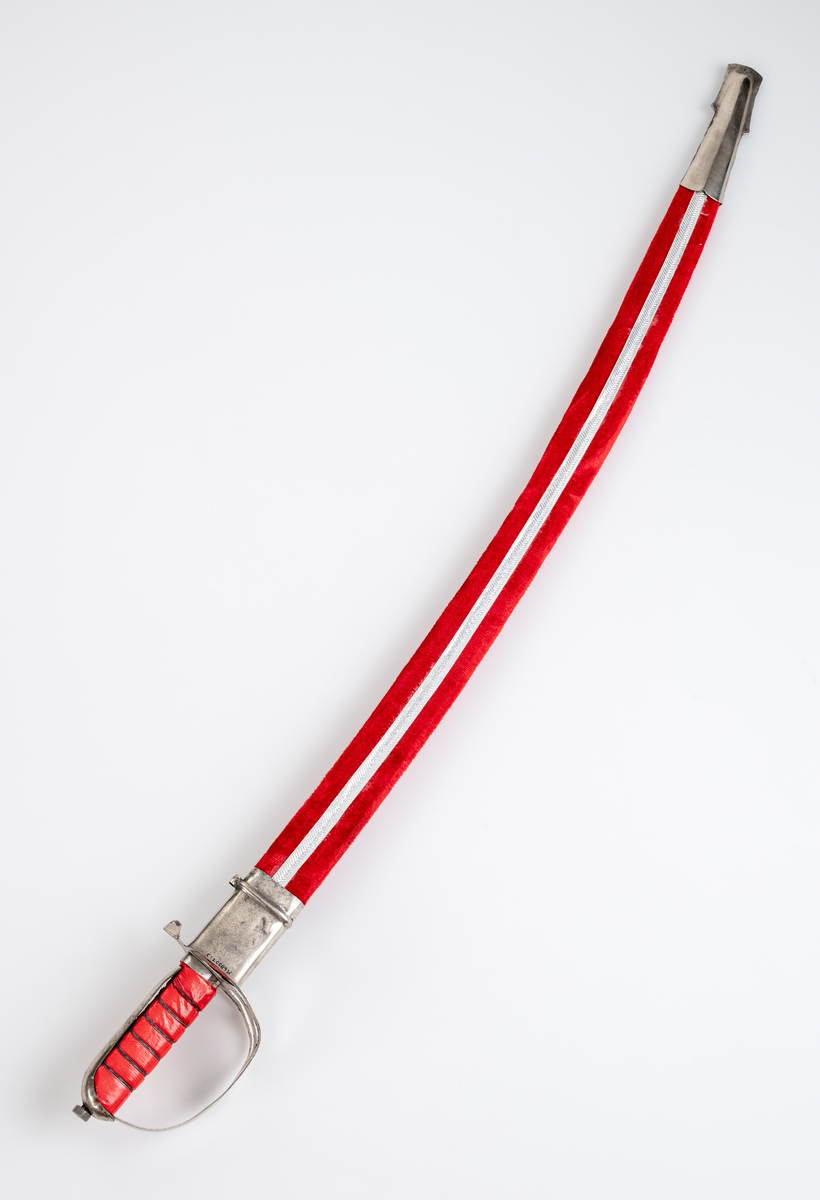 Sabel med balja. Souvenir. Kavel (trä) klädd med röd plastfolie och tvinnad ståltråd som dekor. Parerplåt i aluminium. Klinga av stål med etsad dekor. Balja i läder, klädd med röd plysch och silverfärgat dekorband. Munbleck och släpsko i aluminiumplåt.