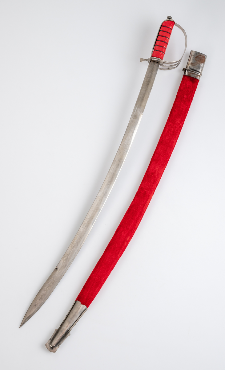 Sabel med balja. Souvenir. Kavel (trä) klädd med röd plastfolie och tvinnad ståltråd som dekor. Parerplåt i aluminium. Klinga av stål med etsad dekor. Balja i läder, klädd med röd plysch och silverfärgat dekorband. Munbleck och släpsko i aluminiumplåt.