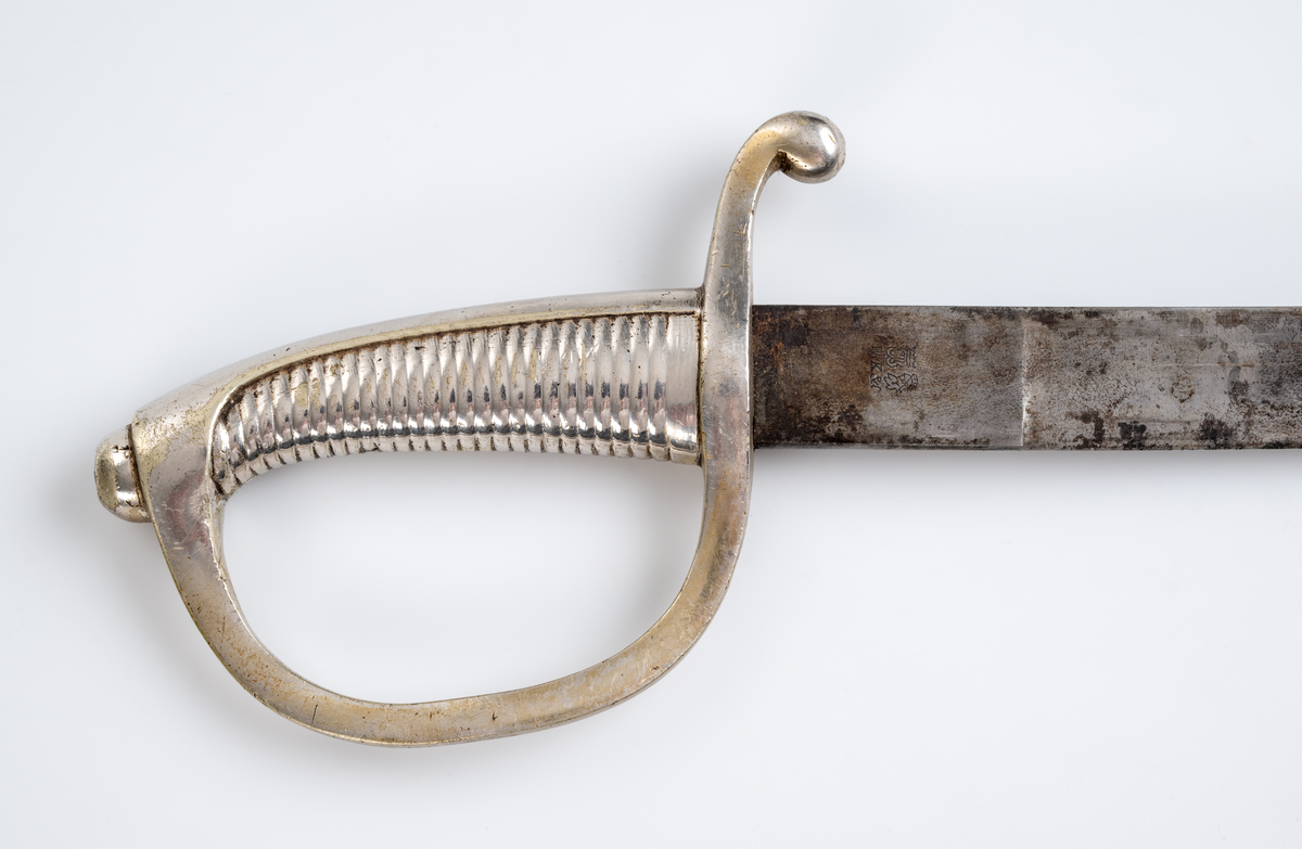 Sabel med balja. Fäste och parerplåt i förnicklad mässing. Klinga av stål. Balja i brunt läder med munbleck och släpsko i förnicklad mässing. Osäkert om baljan ursprungligen hör till sabeln.