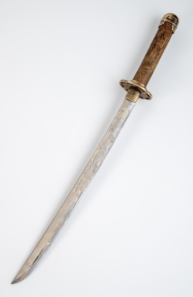 Kortsvärd. Svagt böjt, eneggat blad. Präglad dekor längs med bladets båda sidor. Grepp i trä med skuren ornamentik och en drake i relief. Ornamenterad och förgylld parerstång och knapp.