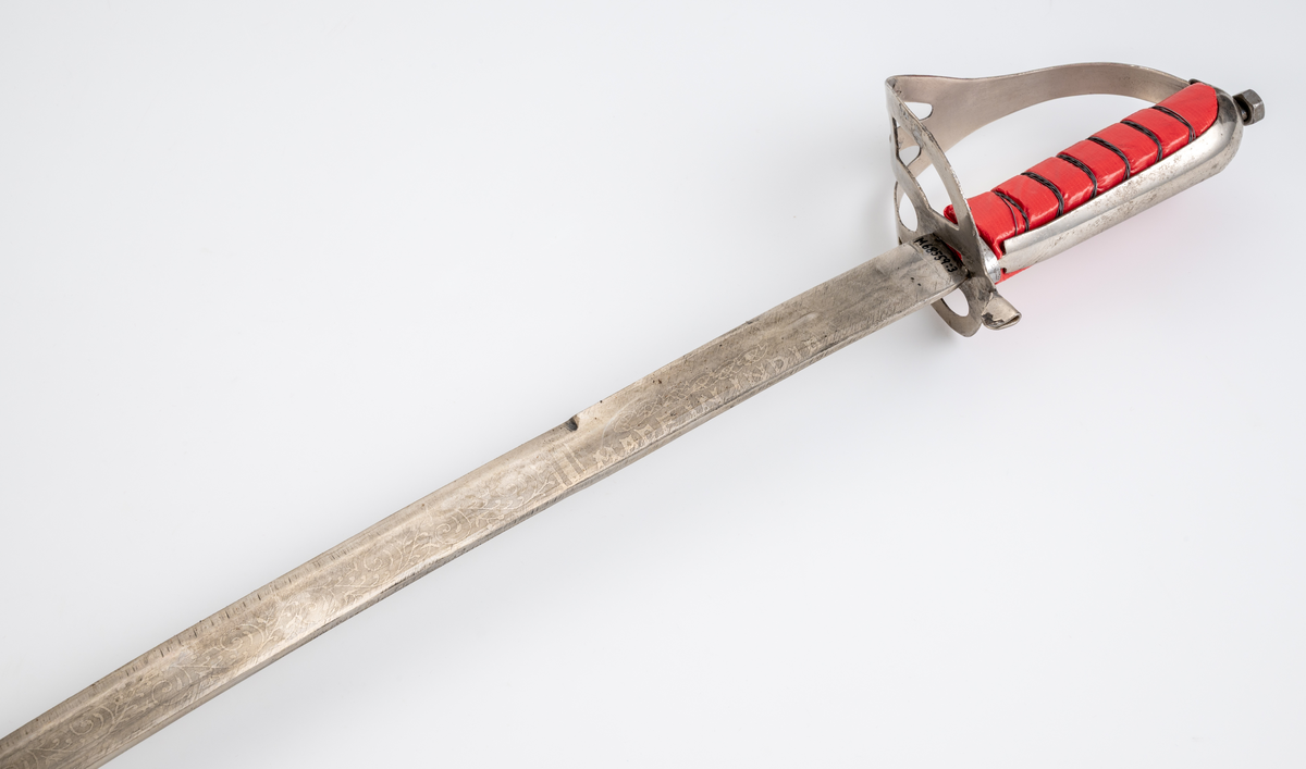 Sabel med balja. Souvenir. Kavel (trä) klädd med röd plastfolie och tvinnad ståltråd som dekor. Parerplåt i aluminium. Klinga av stål med etsad dekor. Balja i läder, klädd med röd plysch och silverfärgat dekorband. Munbleck och släpsko i aluminiumplåt.