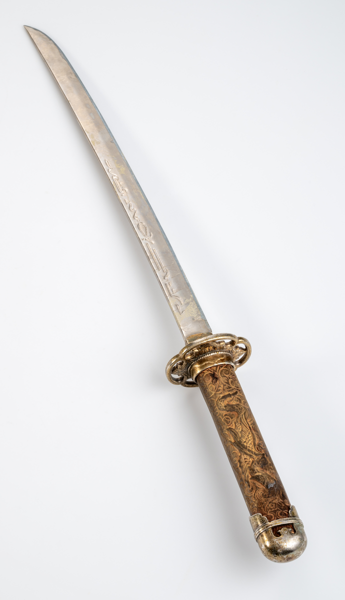 Kortsvärd. Svagt böjt, eneggat blad. Präglad dekor längs med bladets båda sidor. Grepp i trä med skuren ornamentik och en drake i relief. Ornamenterad och förgylld parerstång och knapp.