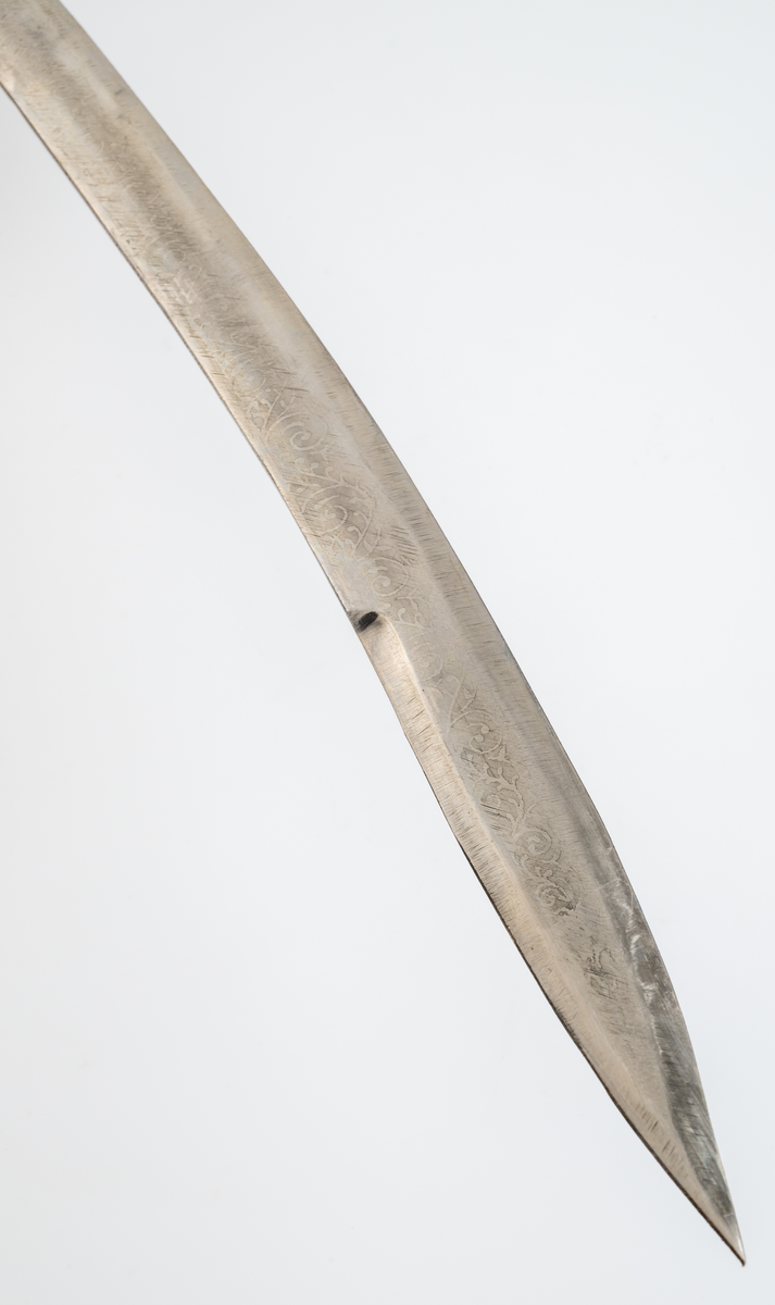 Sabel med balja. Souvenir. Kavel (trä) klädd med röd plastfolie och tvinnad ståltråd som dekor. Parerplåt i aluminium. Klinga av stål med etsad dekor. Balja i läder, klädd med röd plysch och silverfärgat dekorband. Munbleck och släpsko i aluminiumplåt.