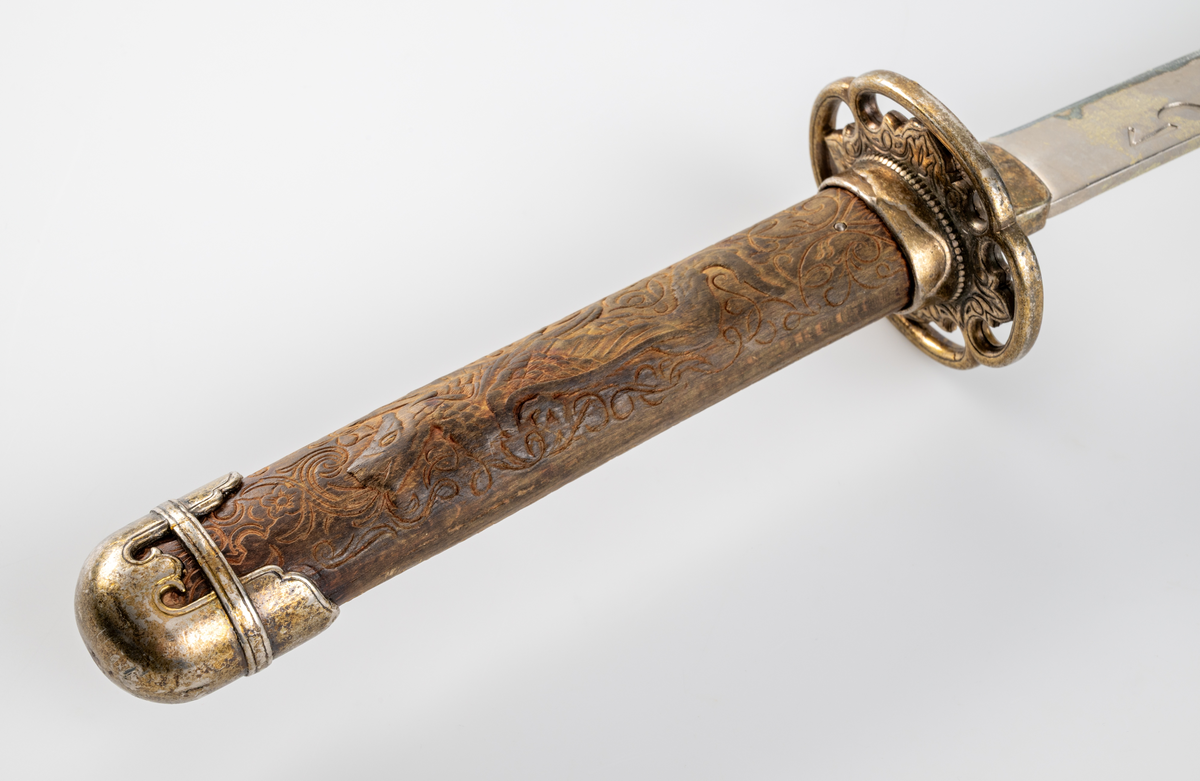 Kortsvärd. Svagt böjt, eneggat blad. Präglad dekor längs med bladets båda sidor. Grepp i trä med skuren ornamentik och en drake i relief. Ornamenterad och förgylld parerstång och knapp.