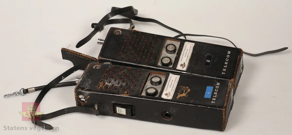 2 stk Walkie Talkie av plast og metall, sølv og svart farget. De er batteridrevet med uttak for 12V drift. Ligger i svarte lærhylstre. Merket med Vegdirektoratet. Lærreim for å bærbarhet. 