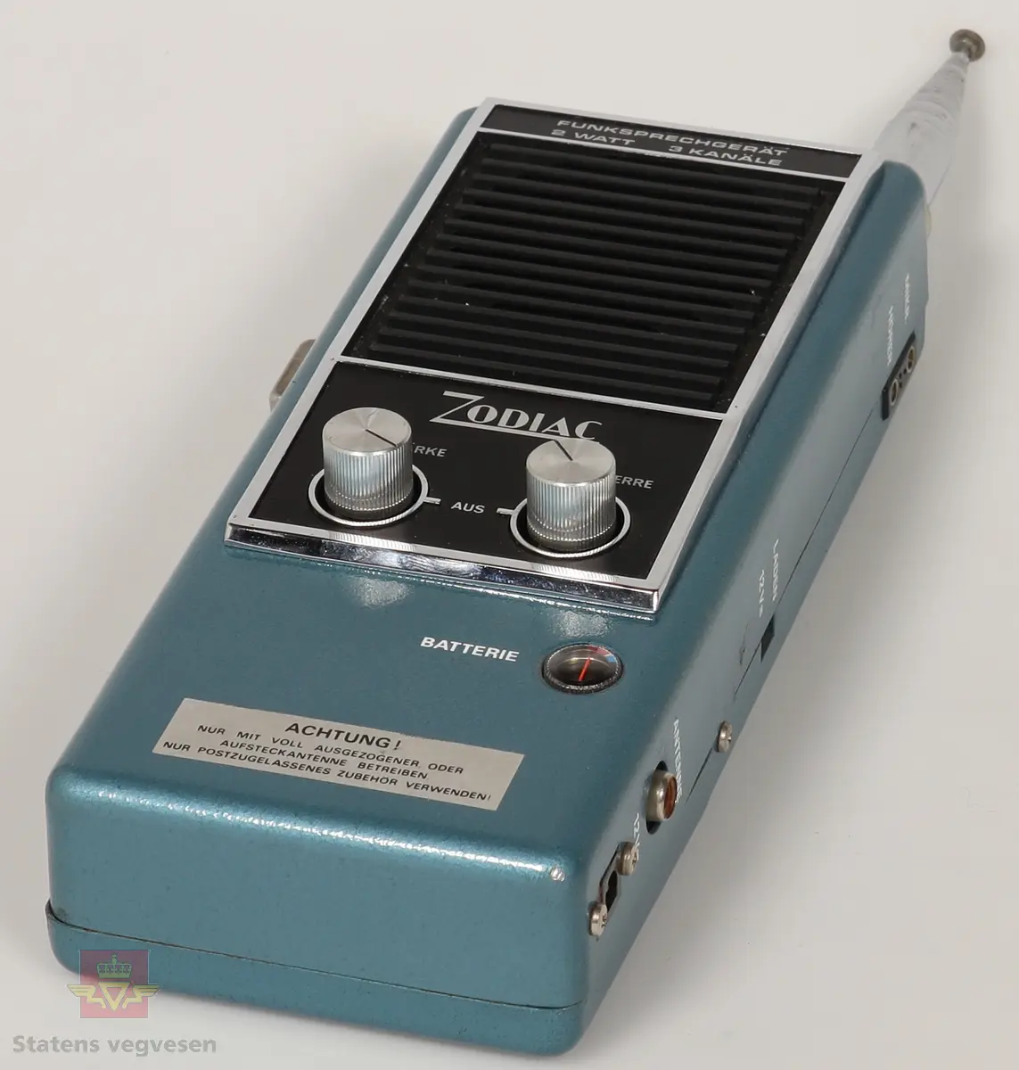 Svart, blå og blank Walkie Talkier. Drives av 8 stk AA batterier. Disse er fjernet. Den har tre kanaler. Skriften på gjenstanden er på tysk. Walkie Talkie ligger i svart skinnveske med glidelås og skinnreim. 