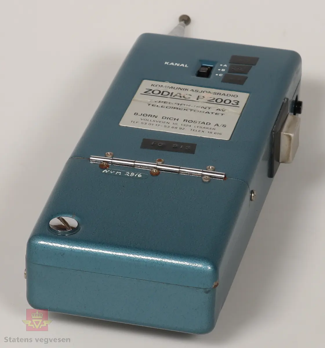 Svart, blå og blank Walkie Talkier. Drives av 8 stk AA batterier. Disse er fjernet. Den har tre kanaler. Skriften på gjenstanden er på tysk. Walkie Talkie ligger i svart skinnveske med glidelås og skinnreim. 
