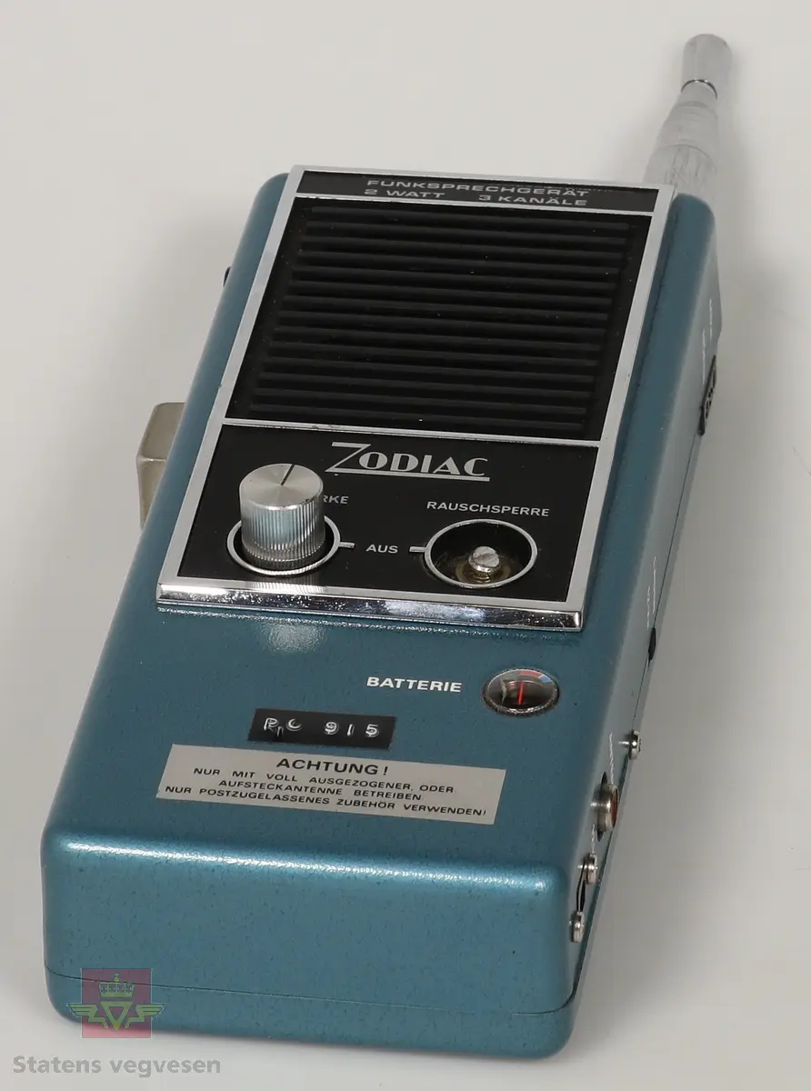 Svart, blå og blank Walkie Talkier. Drives av 8 stk AA batterier. Disse er fjernet. Den har tre kanaler. Skriften på gjenstanden er på tysk. Mangler en knapp. Defekt. Walkie Talkie ligger i svart skinnveske med glidelås og skinnreim. 