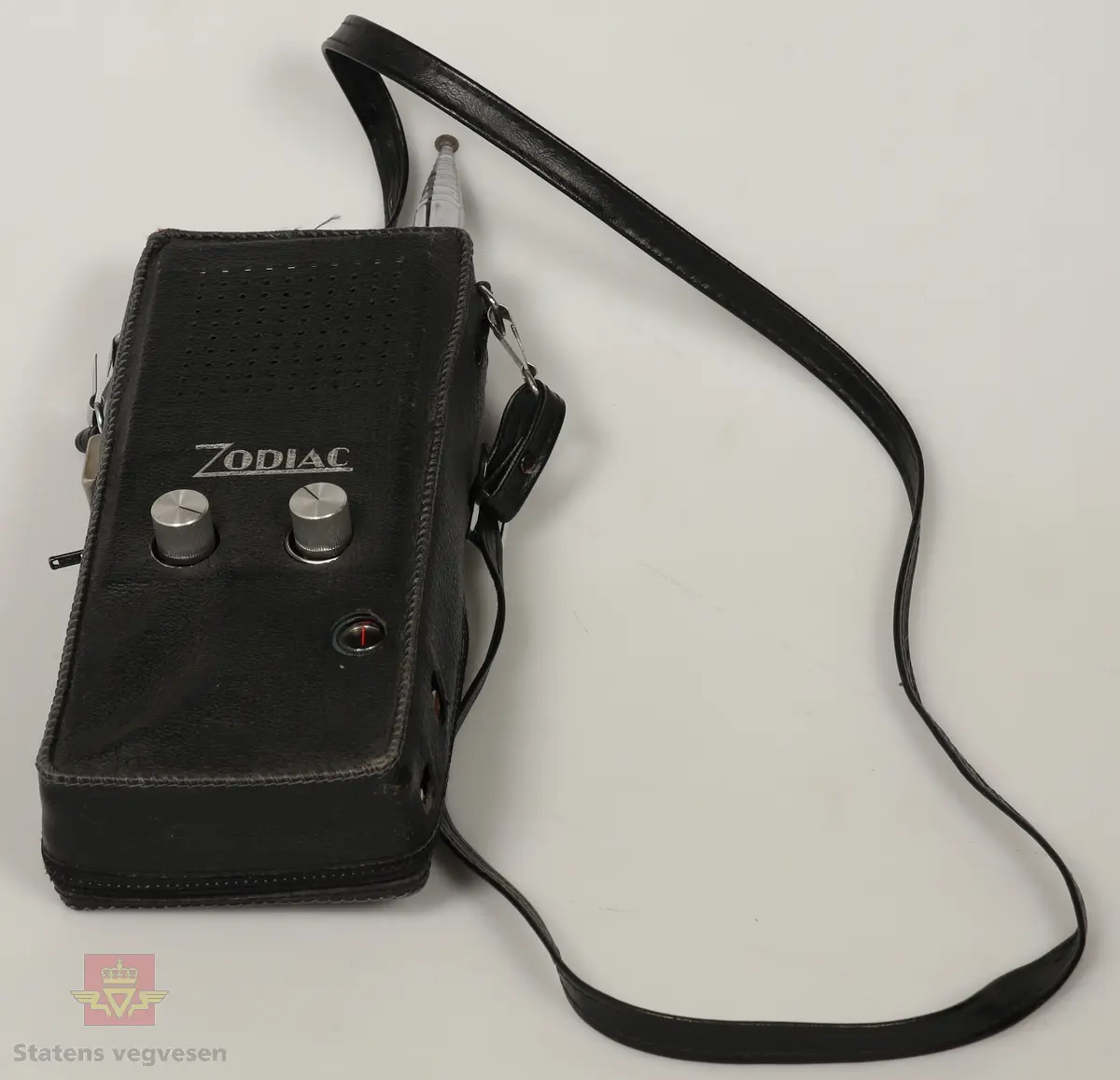 Svart, blå og blank Walkie Talkier. Drives av 8 stk AA batterier. Disse er fjernet. Den har tre kanaler. Skriften på gjenstanden er på tysk. Walkie Talkie ligger i svart skinnveske med glidelås og skinnreim. 
