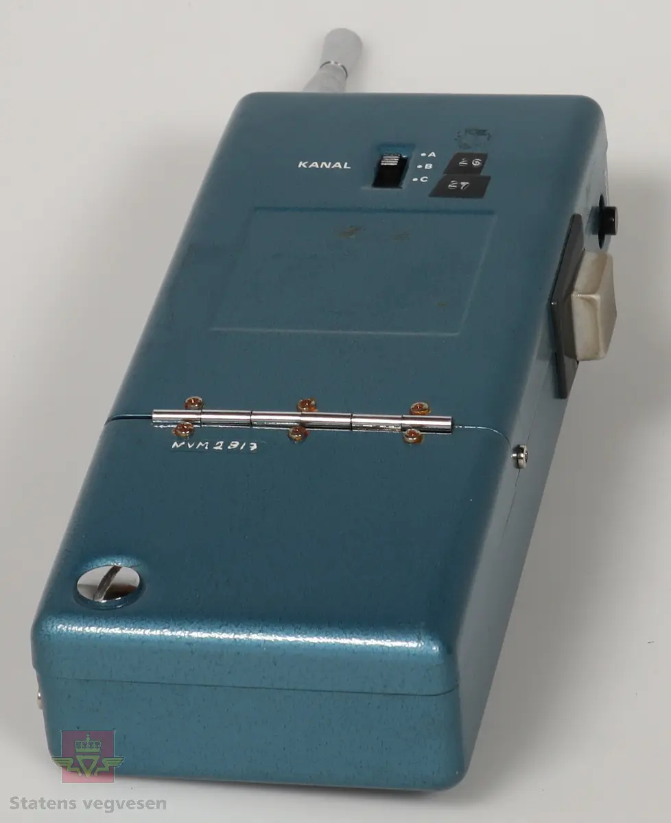 Svart, blå og blank Walkie Talkier. Drives av 8 stk AA batterier. Disse er fjernet. Den har tre kanaler. Skriften på gjenstanden er på tysk. Mangler en knapp. Defekt. Walkie Talkie ligger i svart skinnveske med glidelås og skinnreim. 