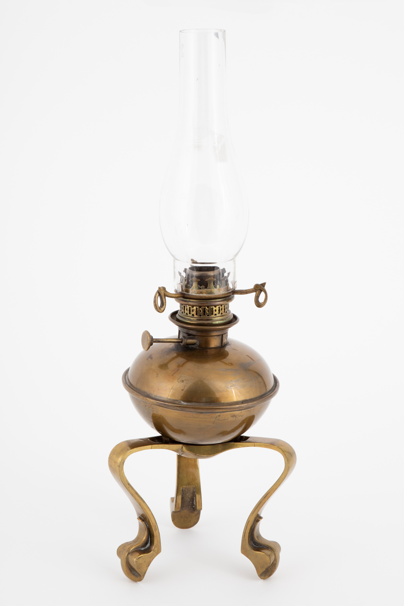 Oljelampe i drevet og støpt messing, og med kuppel i glass. Messingen har fått en grønnlig tone ved en halvmatt ferniss. Øvre del har en lett flattrykt kuleform, hvortil festet en holder med skruer for lampeglass og kuppel. Kulen står løst med støttetapper på den støpte foten. Foten har tre kraftige ben med svungne former.