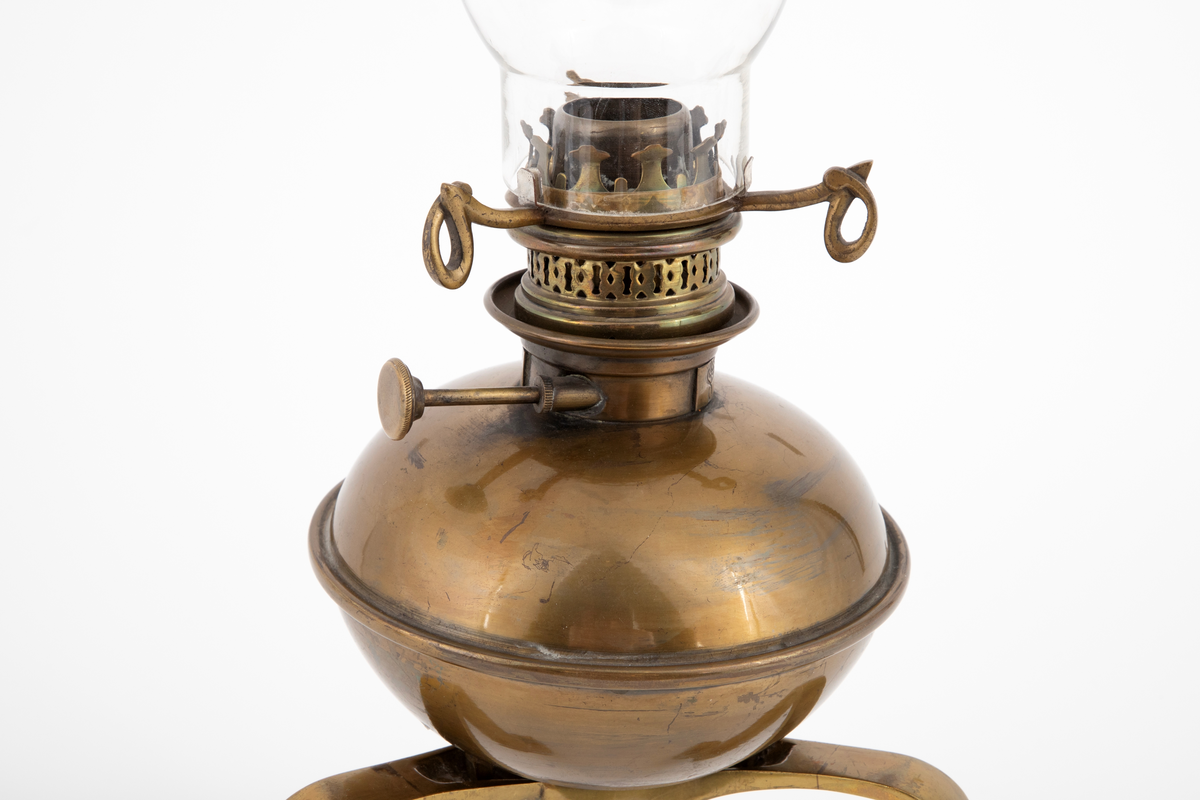 Oljelampe i drevet og støpt messing, og med kuppel i glass. Messingen har fått en grønnlig tone ved en halvmatt ferniss. Øvre del har en lett flattrykt kuleform, hvortil festet en holder med skruer for lampeglass og kuppel. Kulen står løst med støttetapper på den støpte foten. Foten har tre kraftige ben med svungne former.