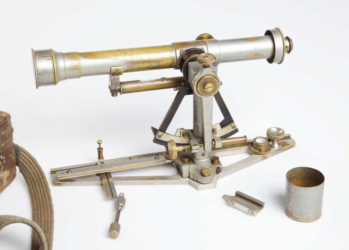 Mått: inst. tuben är 29,8 cm och ca 16,7 cm hög. Trpt. lådan är 33,5 x 13,5 x 20 cm.

Övriga uppgifter: optisk mätningsinstrument. Bestående av en linsförsedd mässingtub fäst på ett förnicklat mässingsstativ. Stativet har mätskalor samt två vattenpass. Ett är tubformat med två skalfönster, det andra, längre ner på stativets fot, är cirkeltunt och platt. 
Tillhörande transportlåda av trä med sinkade hörn, locket låses med två mässingshaspar. Sekundär bärrem av textil. 

Hela instumentet har vid användning varit placerat på ett stativförsett mätbord. 
Funktion: Avvägning och kartering av torrläggningsföretag