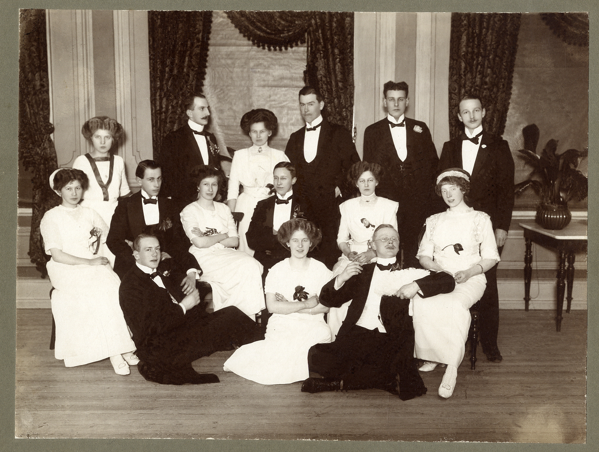 Skottårsbal i "Stadts" pelarsal, Växjö 1912.
En grupp ungdomar har samlats för gruppfoto.
Text på baksidan:
"Skottårs bal i "Stadts" pelarsal 1912. Fr. höger, övre raden: Rudolf Feuk, Gustaf Tausis, Julius Lundberg, Ester Lundberg, f. Feuk, Karl Wahlström, Clara Friman, f. Johnsson. 
Andra raden, sittande: Vivan Feuk, f. Andersson , Ellen Tausis, f. Johansson, Yngve Ahlsson (?), Alma Tollin (?), f. Sposselin (?), Harry Friman, Betty Hvitfeldt (eg. Widfeldt), f. Hallenborg.
På golvet: Karl Boberg (?), Hilma Toll, f. Pettersson, Folke Sundberg."