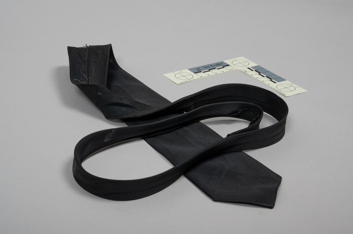 Slips i svart. Bak på slipset står det følgende på fabrikatmerket: "Made from Terylene". Slipset var hengt på skuldren på vesten med reg.nr. LSO 2325 (STMU-009006).