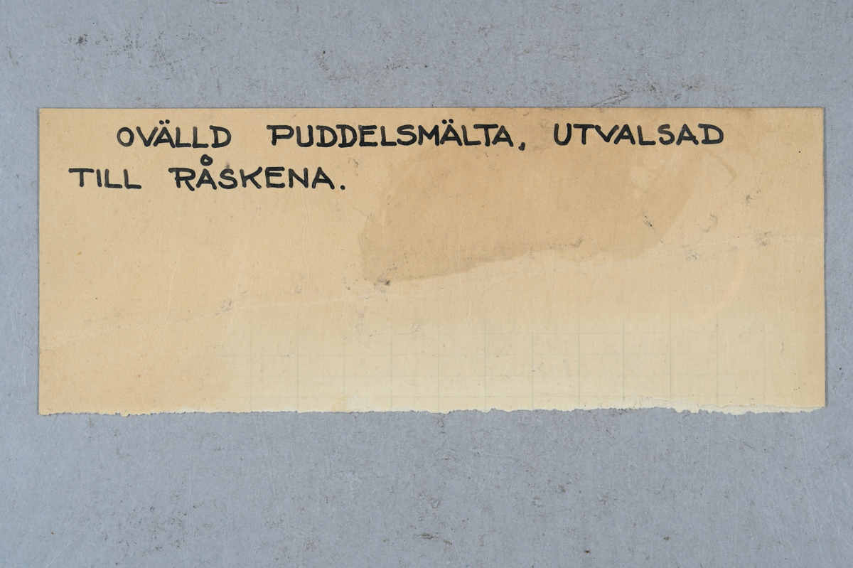 Prov på ovälld puddelsmälta. Påklistrad lapp på provet med notering: "Owälld puddlingssmälta af tackjern. OL (?) ...(?). 82° (?) Puddlingsugnen M...(?) ...(?). Nyby gå..(?) 1838 (?)." Tillhörande lapp med notering: "Ovälld puddelsmälta, utvalsad till råskena."