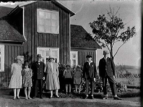 Åtta finklädda personer framför en byggnad, samma som på bildnr E7407-7408. Fyra pojkar och fyra flickor, alla i olika huvudbonader. Bilden troligen tagen i Gällared eller Okome.