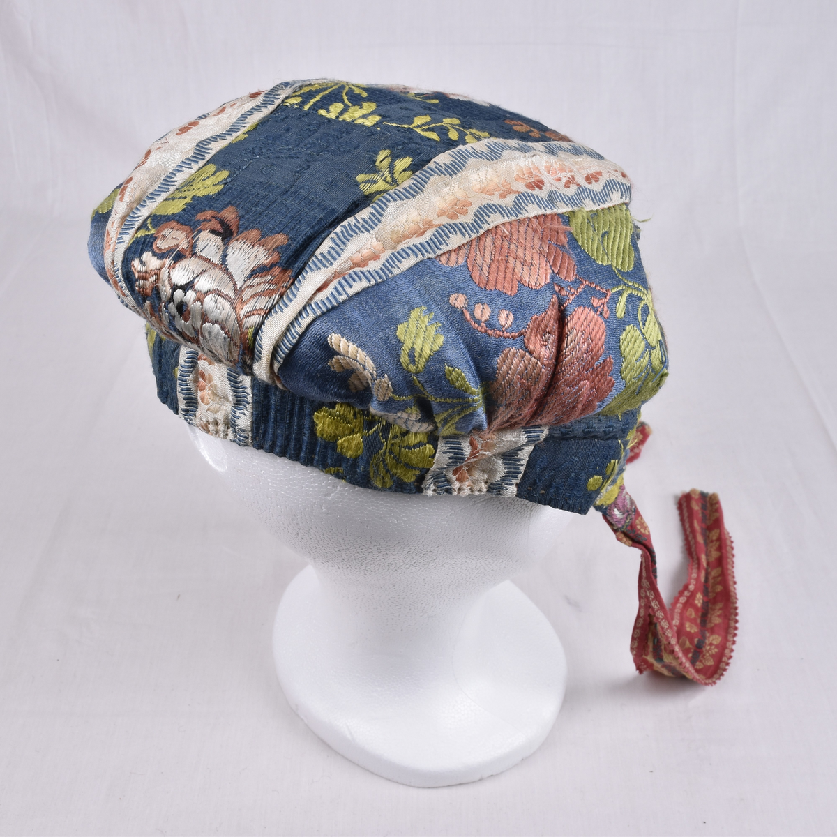 Rynkelue i mønstervevd silke. Lua er satt sammen av mange små biter, og det er flere ulike typer stoff i den. Fôret med linstoff. Den rundede pullen er rynket og sydd til et smalt pannestykke som går over panna og ender i et knytebånd på hver side.