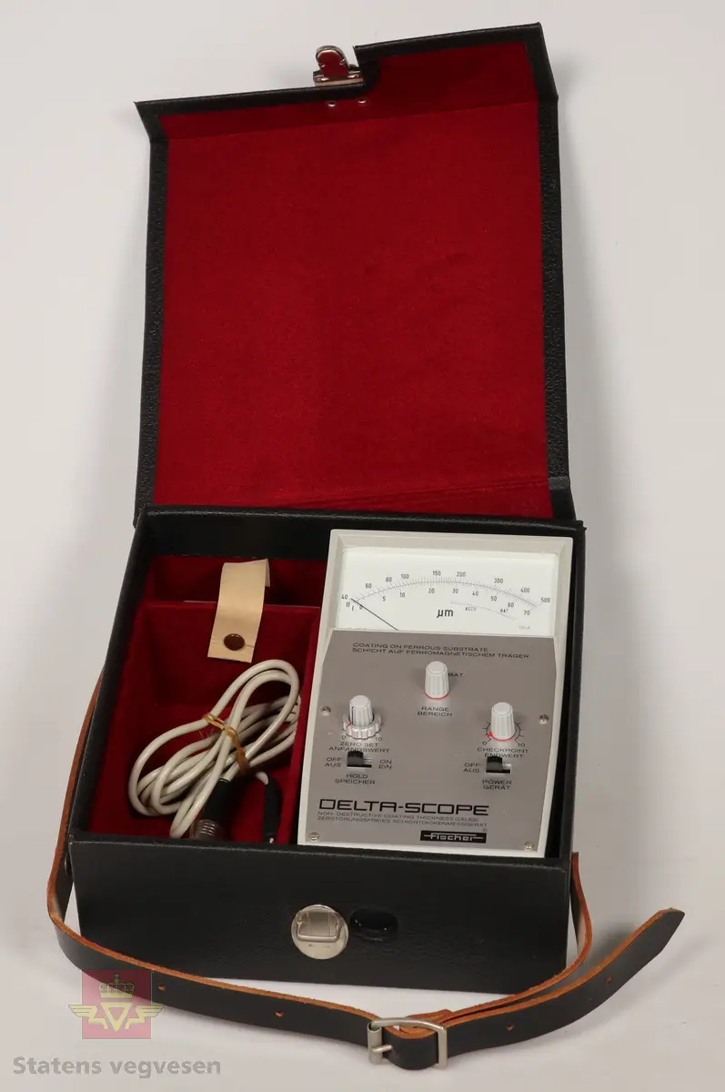 Måleinstrument laget av plast og aluminium. Gråfarget med hvit viser skjerm, svart tekst og grå knotter. Ligger i svart koffert med skinnreim og rødt fløyelsfor. Tilbehør er batteri, type tørr, Varta Spesial 28, 9 Volt. Flerfarget. Og føler til my-måler som har grå ledning og svarte og blanke kontakter. 