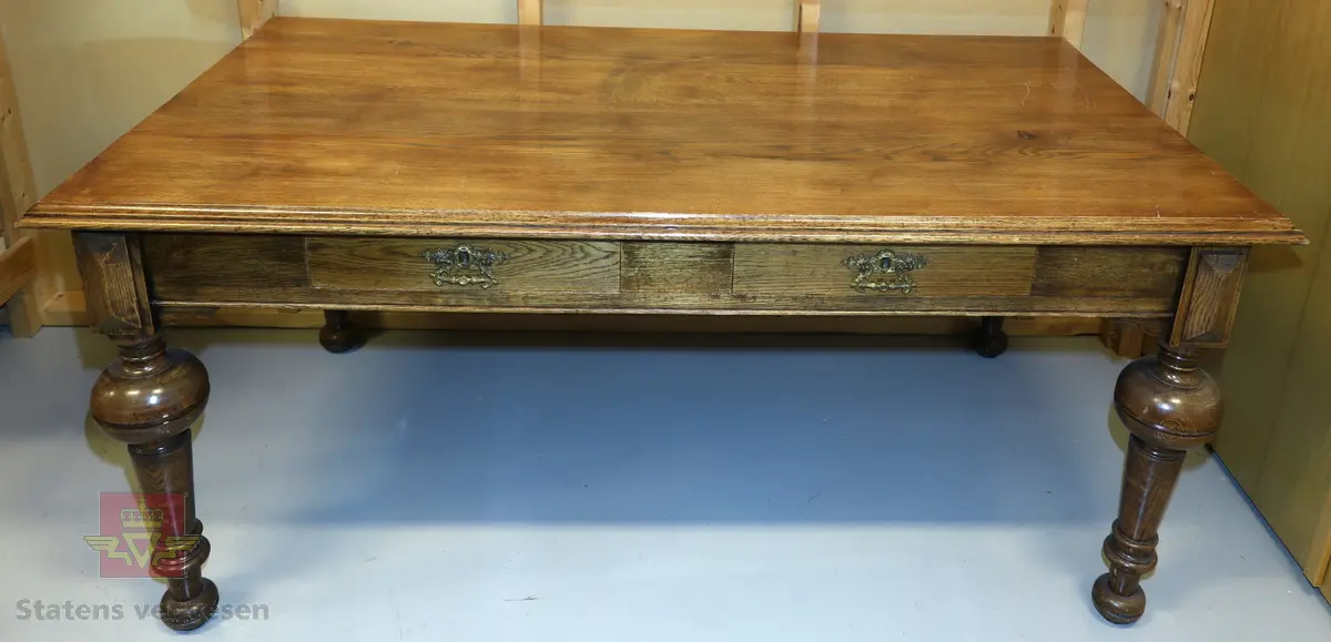 Brunt solid skrivebord av treverk med to uttrekksskuffer. Bena er dreid, og det er fine utsmykninger i hjørner og langs bordkanten. Skuffer har lås og messinghåndtak. En del riper på bordplate.