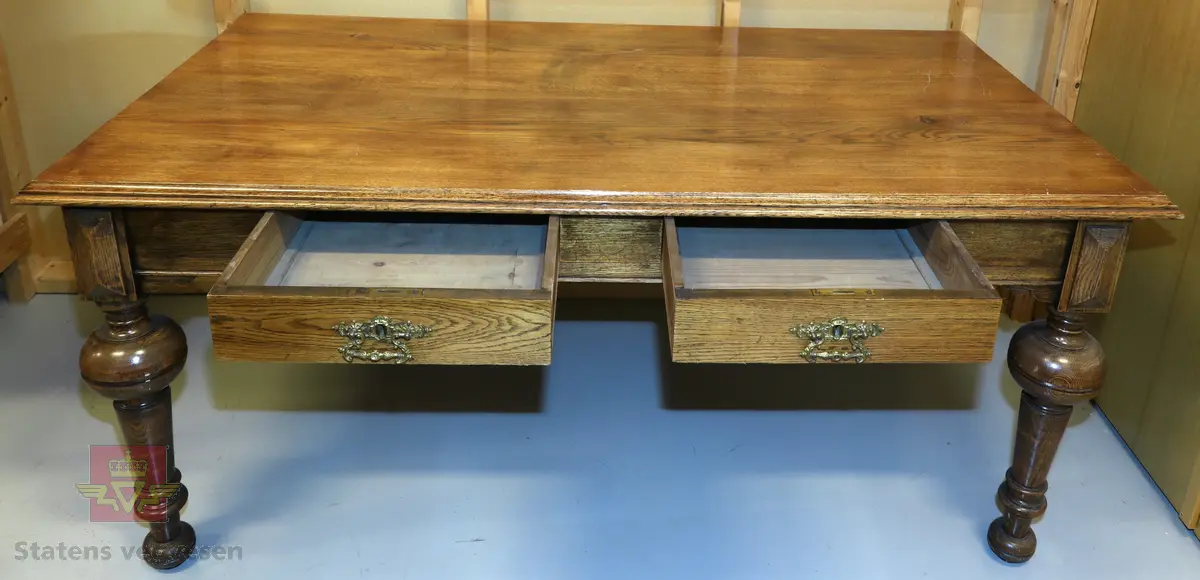Brunt solid skrivebord av treverk med to uttrekksskuffer. Bena er dreid, og det er fine utsmykninger i hjørner og langs bordkanten. Skuffer har lås og messinghåndtak. En del riper på bordplate.