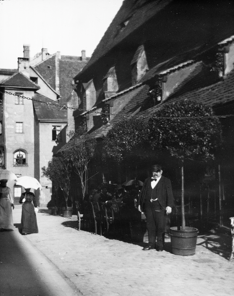 Skioptikonbild med motiv man framför serveringen Bratwurst-Glöcklein vid Moritzkapellet i Nürnberg. Bredvid promenerar två kvinnor med paraply.
Bilden har förvarats i kartong märkt: Nürnberg VI. 1901