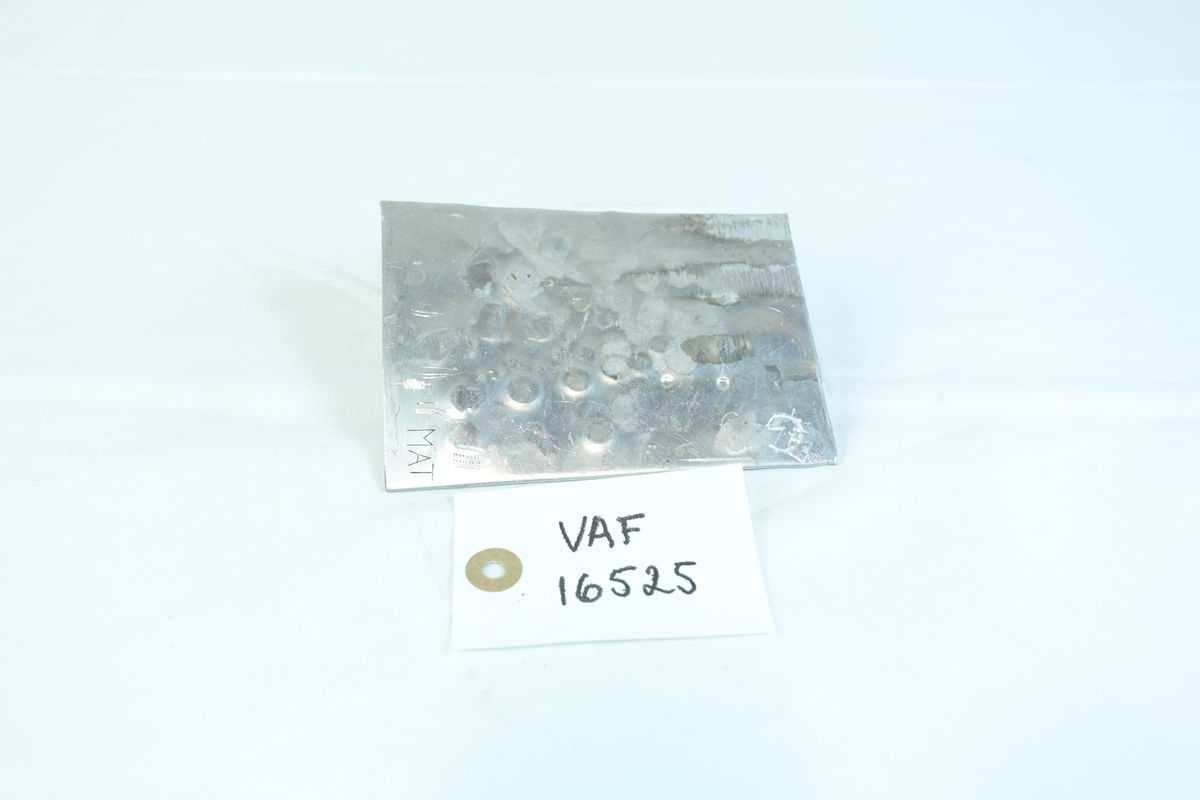 Firkantformet metallflak med sirkelformede merker og bokstavene M, A og T i et hjørne. Trolig brukt til materiale i gullsmedvirksomhet.