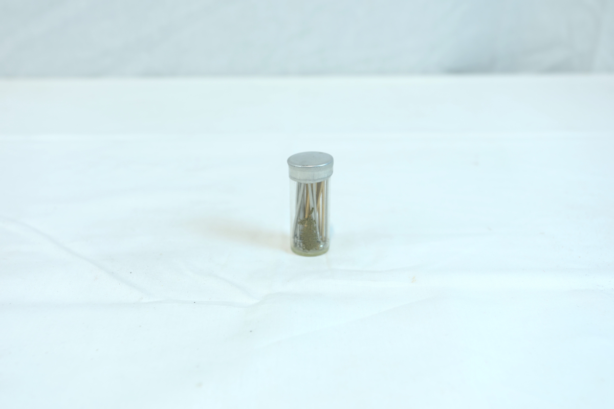 Emballasjeglass med lokk. I glasset er det tynne avlange metallgjenstander. Trolig materiale eller verktøy i gullsmedvirksomhet.