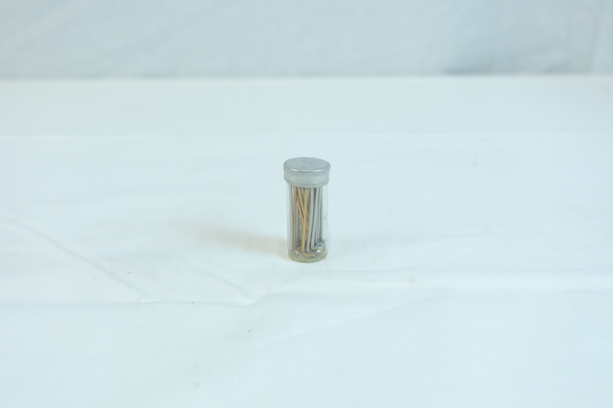 Emballasjeglass med lokk. I glasset er det tynne avlange metallgjenstander. Trolig materiale eller verktøy i gullsmedvirksomhet.
