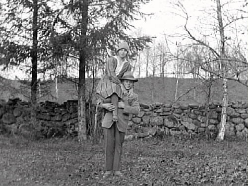 Syskonen Gertrud Torsson och Ragnar Torsson i Skog, Gällared. Gertrud sitter på Ragnars ena axel. De står framför en stengärdsgård och i bakgrunden ses en skogsbacke med glesa träd.