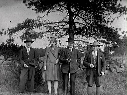 Tre män och en kvinna står vid en stengärdsgård intill en tall. Mannen i mitten hette Nores Karlsson och var gårdfarihandlare. Han brukade bo hos Nora som var ensamstående och bodde i ett torp under Klockaregården i Gällared. Noras fosterdotter Svea Svärd står bredvid Nores. På ytterkanterna står bröderna Nordenskjöld, som uppvaktade Svea.