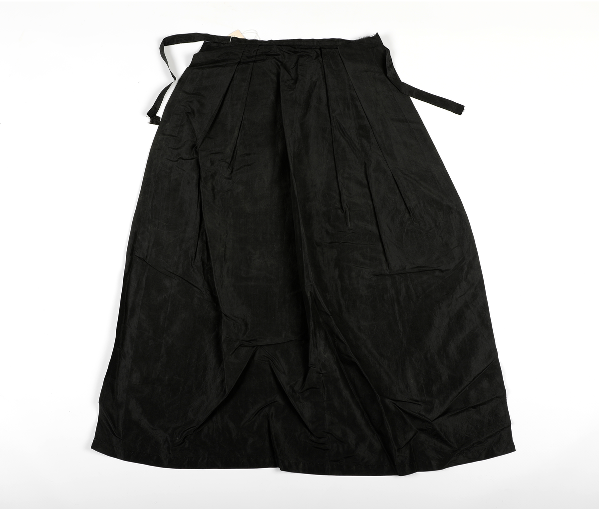 Sort forkle i tynt, sort stoff, med sløyfebånd til knyting, som er en forlengelse av øvre kant på forkleet. 