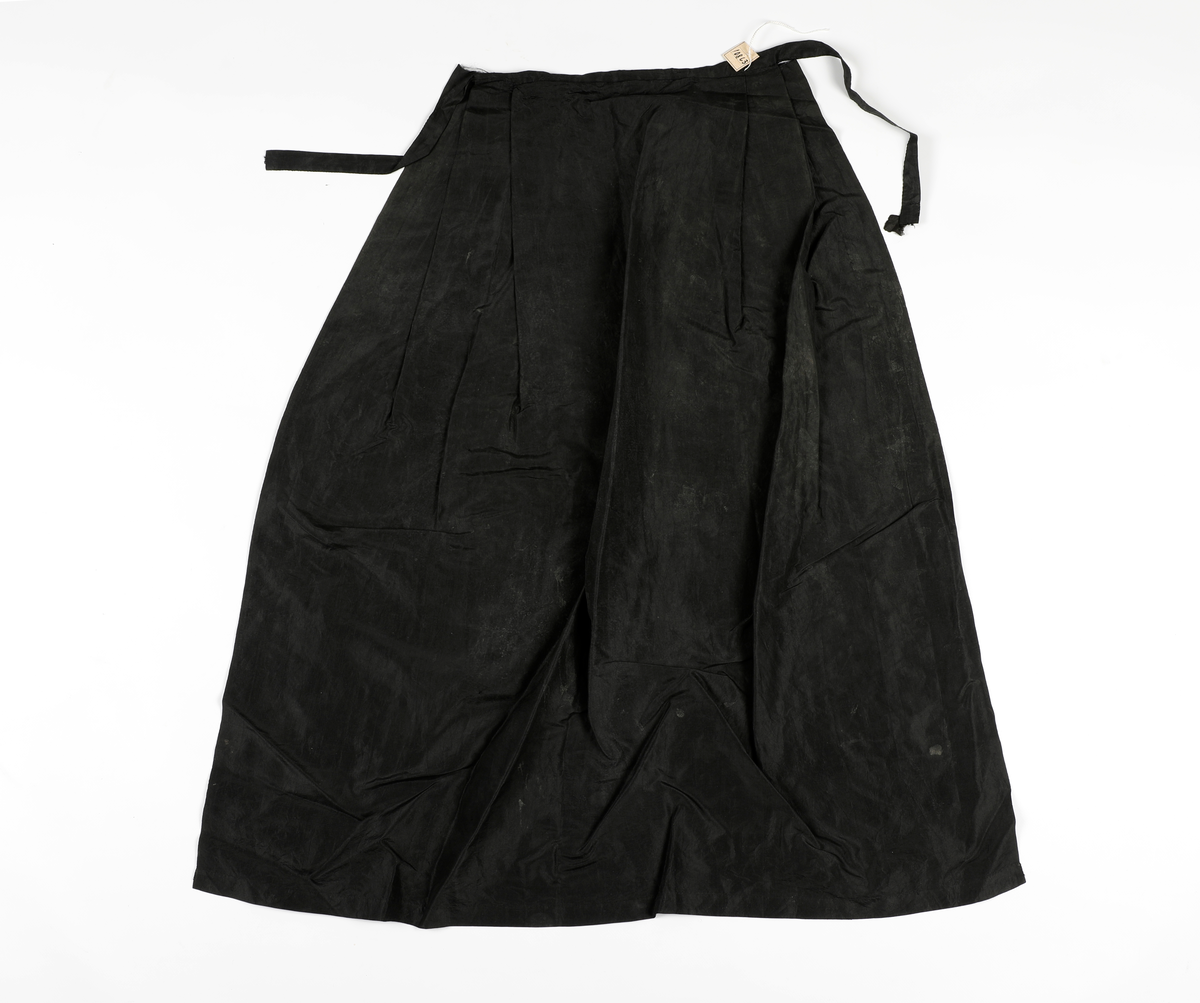 Sort forkle i tynt, sort stoff, med sløyfebånd til knyting, som er en forlengelse av øvre kant på forkleet. 