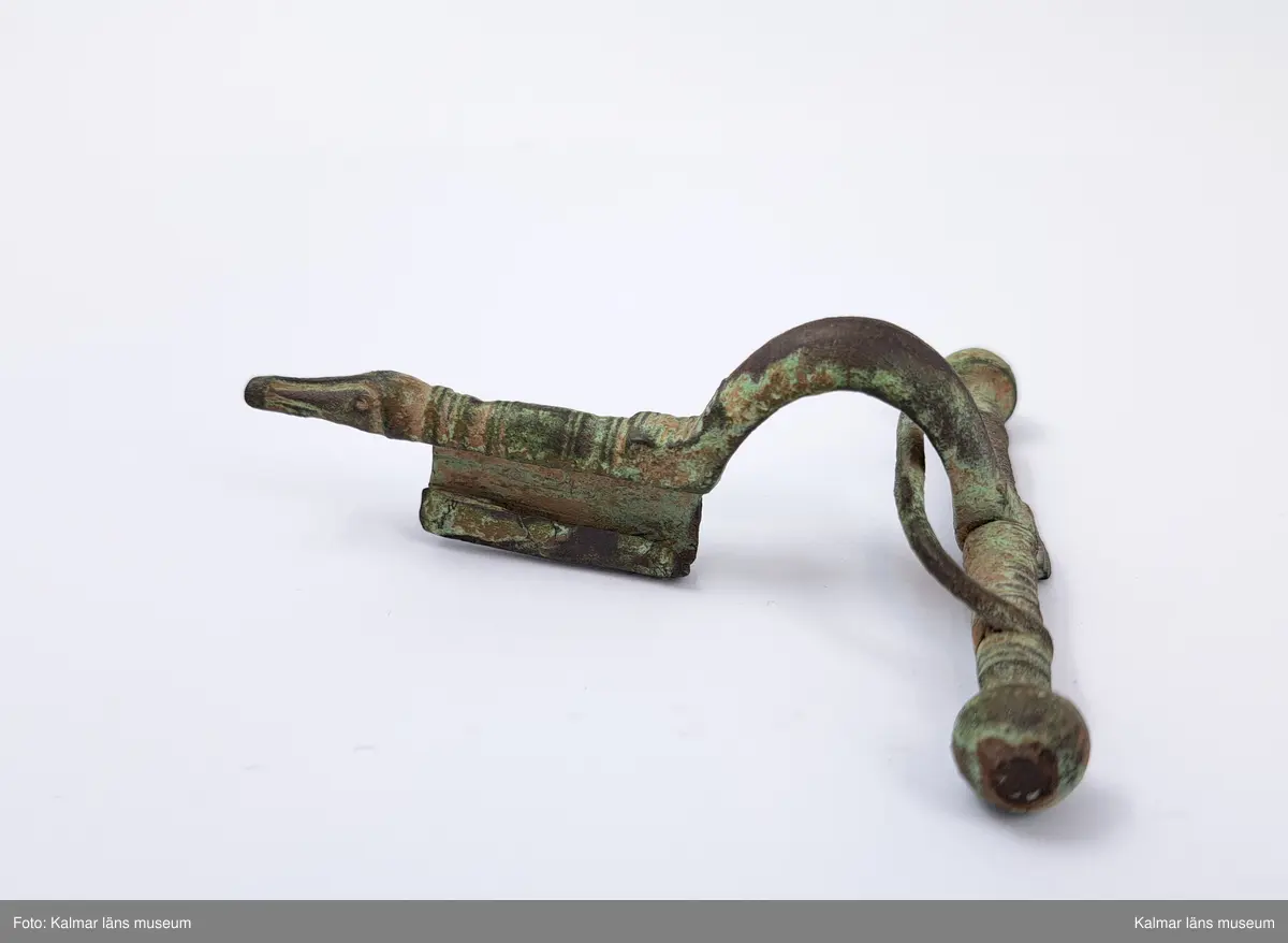 KLM 1458. Fibula, armborstfibula. Gjuten av brons. Fotstycket avslutas med ett djurhuvud och nålhållaren undertill är helt öppen. Själva nålen saknas. Tvärstång med ändknoppar.