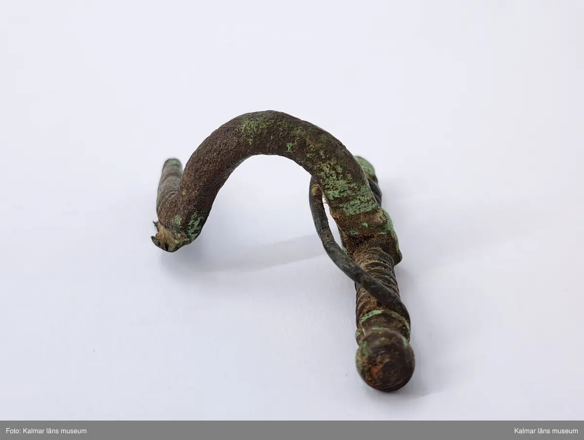 KLM 1248. Fibula, armborstfibula. Fotstycket slutar tvärt nedtill. Nålhållaren är böjd vinkelrätt åt sidan. Tvärstång med olikformade knoppar.