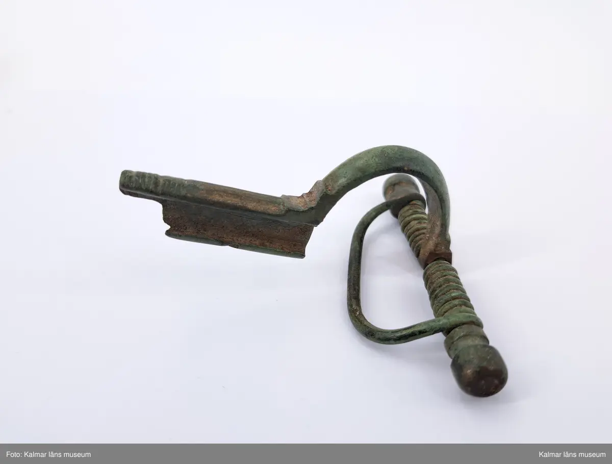 KLM 117. Fibula, armborstfibula. Gjuten av brons. Fotstycket har en tvär avslutning och är nederst prytt med inpunsade tvärlinjer. Den öppna nålhylsan är något skadad. Nålen saknas.
