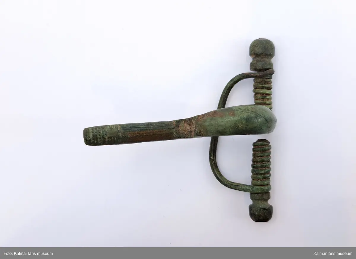 KLM 117. Fibula, armborstfibula. Gjuten av brons. Fotstycket har en tvär avslutning och är nederst prytt med inpunsade tvärlinjer. Den öppna nålhylsan är något skadad. Nålen saknas.